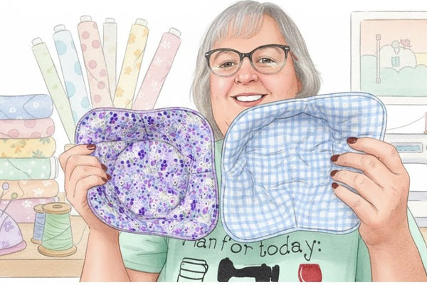 Yvonne holding up two finished quilted bowl cozies, one in a purple floral print and one in blue gingham, in a sewing room