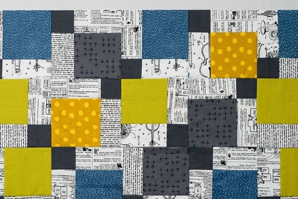 Patchwork quilt top in charcoal, teal, mustard and chartreuse squares set against black and white newsprint print fabric