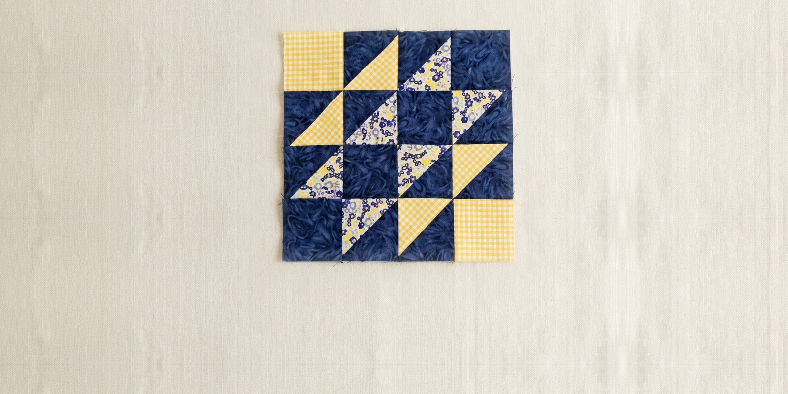 A Hovering Hawks quilt block in navy, yellow gingham and a small floral, sixteen units in a four by four grid with plain light squares in two opposite corners