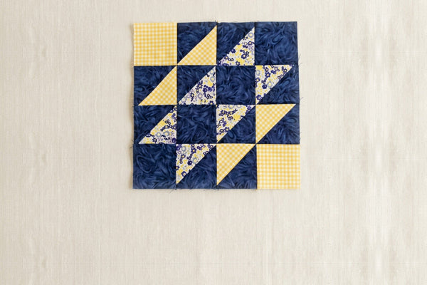 A Hovering Hawks quilt block in navy, yellow gingham and a small floral, sixteen units in a four by four grid with plain light squares in two opposite corners