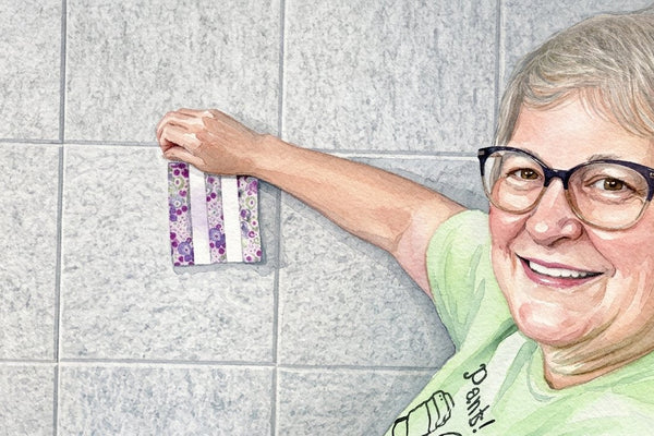 Watercolor illustration of a smiling quilter in glasses holding a small three strip patchwork block flat against a tiled wall