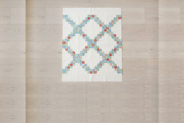 A scrappy double Irish chain quilt top on a pale wood floor, with diagonal chains of small squares in blue, green and red crossing plain cream squares
