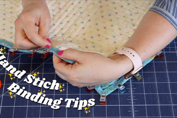 Hands stitching quilt binding down by hand at a mitered corner, with a blue and green scrappy binding