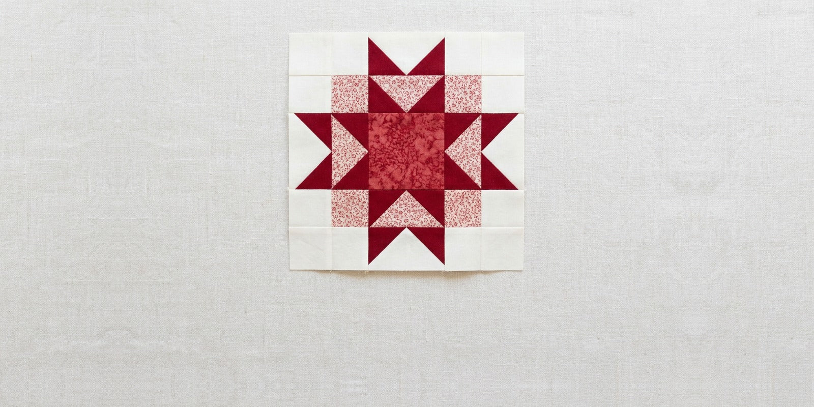 An Ohio star quilt block in deep red and cream on a linen surface, with a pale red print forming a square ring behind the four star points