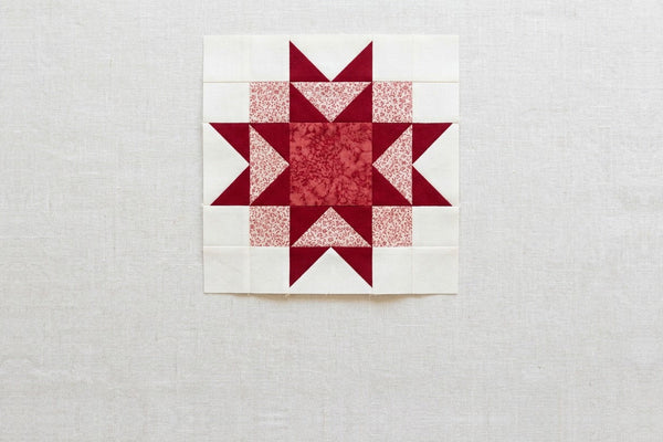 An Ohio star quilt block in deep red and cream on a linen surface, with a pale red print forming a square ring behind the four star points