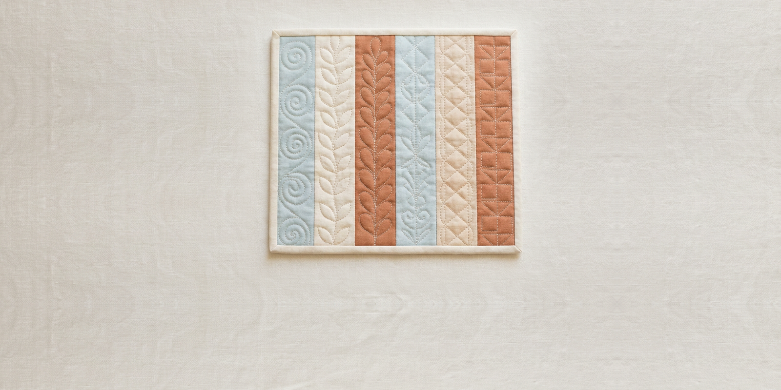 A quilt as you go panel of six fabric strips in blue, cream and terracotta, each strip quilted with a different free motion motif, lying on pale linen