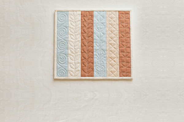 A quilt as you go panel of six fabric strips in blue, cream and terracotta, each strip quilted with a different free motion motif, lying on pale linen