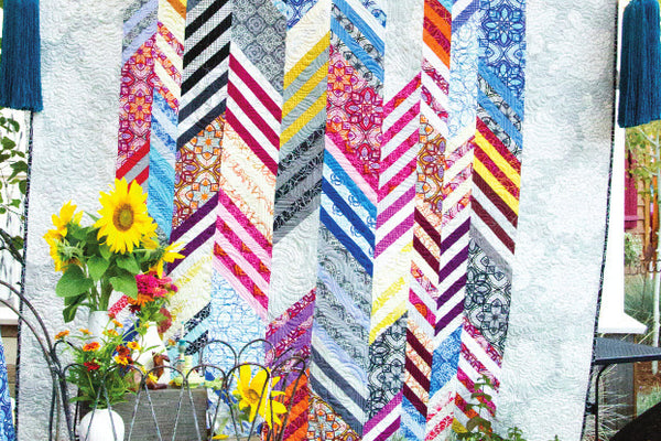 The Souks quilt hanging outdoors, ten vertical rows of angled strips forming chevrons in bright prints against white